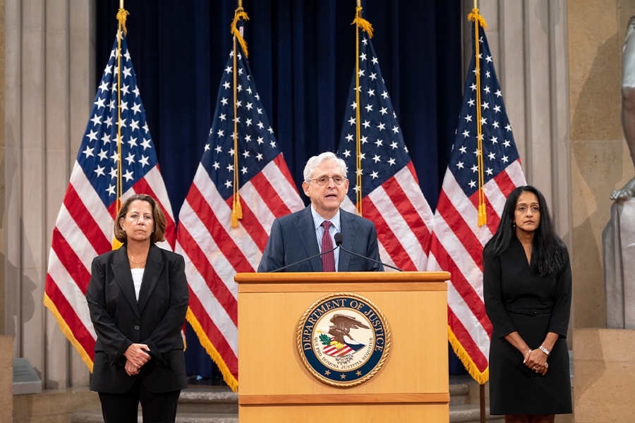 Attorney General Merrick B. Garland delivers remarks from a podium in the Great Hall at the Department of Justice. On his left is Deputy Attorney General Lisa O. Monaco, and on his right is Associate Attorney General Vanita Gupta.