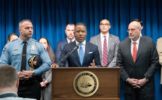 Assistant Attorney General of the Criminal Divison Kenneth A. Polite, Jr. delivers remarks from a podium bearing the United States Attorney's Office for the District of Minnesota seal. To the left stands the Chief of the Minneapolis Police Department Brian O'Hara. To the right stands United States Attorney for the District of Minnesota Andrew M. Luger. Other officials stand in a row behind.