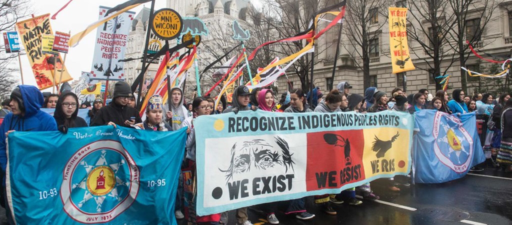 A group of American Indian and Alaska Natives walking down a city street as part of a demonstrating, holding a sign that reads, "Recognize Indigenous People's Rights. We exist. We resist. We rise" 