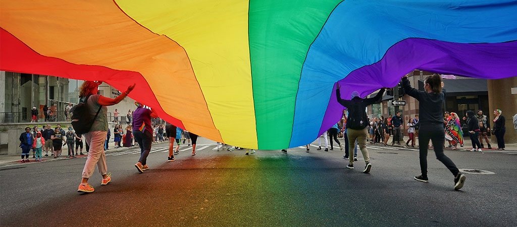 A group of people walk down a street carrying a large LGBTQ pride flag.