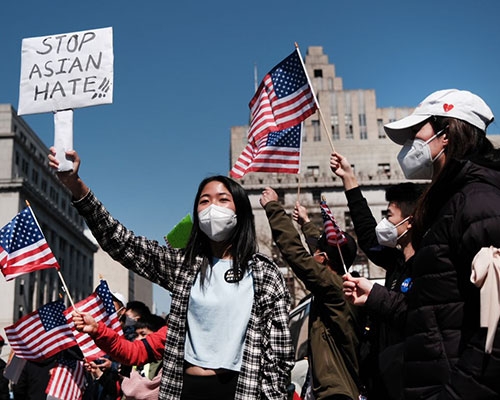An Asian woman holds a sign that reads "Stop Asian Hate" at a demonstration. Protesters in the background hold the American flag. 